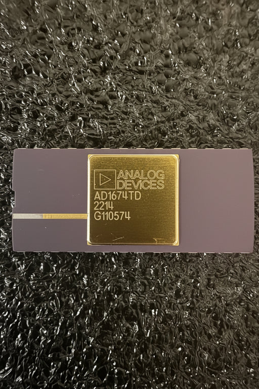 Analog Devices AD1674TD analog-to-digital converter chip in its original dark gray and gold packaging, resting on a textured black surface.